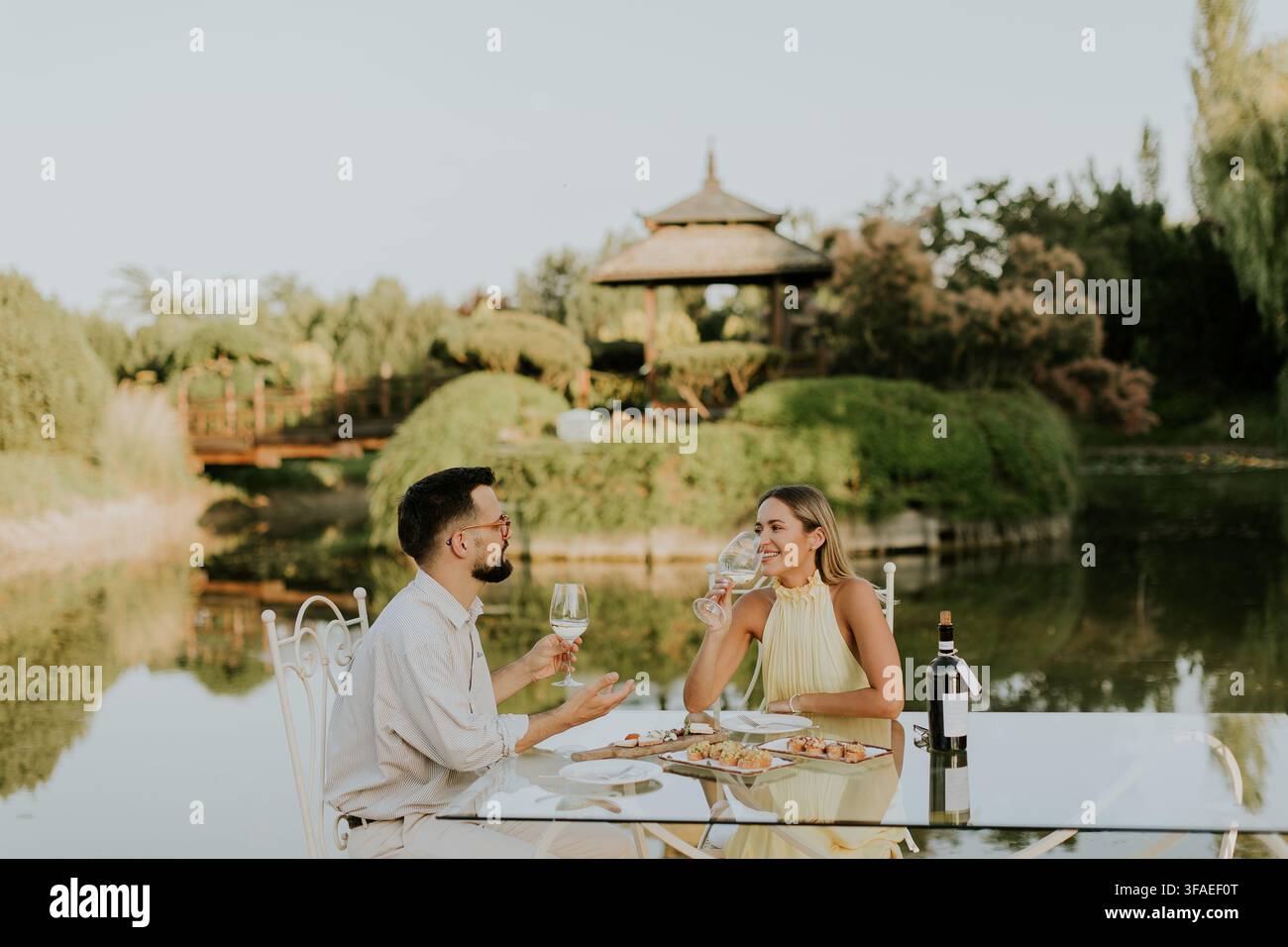 Ein Paar genießt ein intimes Abendessen an einem stilvollen Tisch am Wasser. Sie teilen Gelächter und feines Essen, umgeben von der Schönheit der Natur. Stockfoto