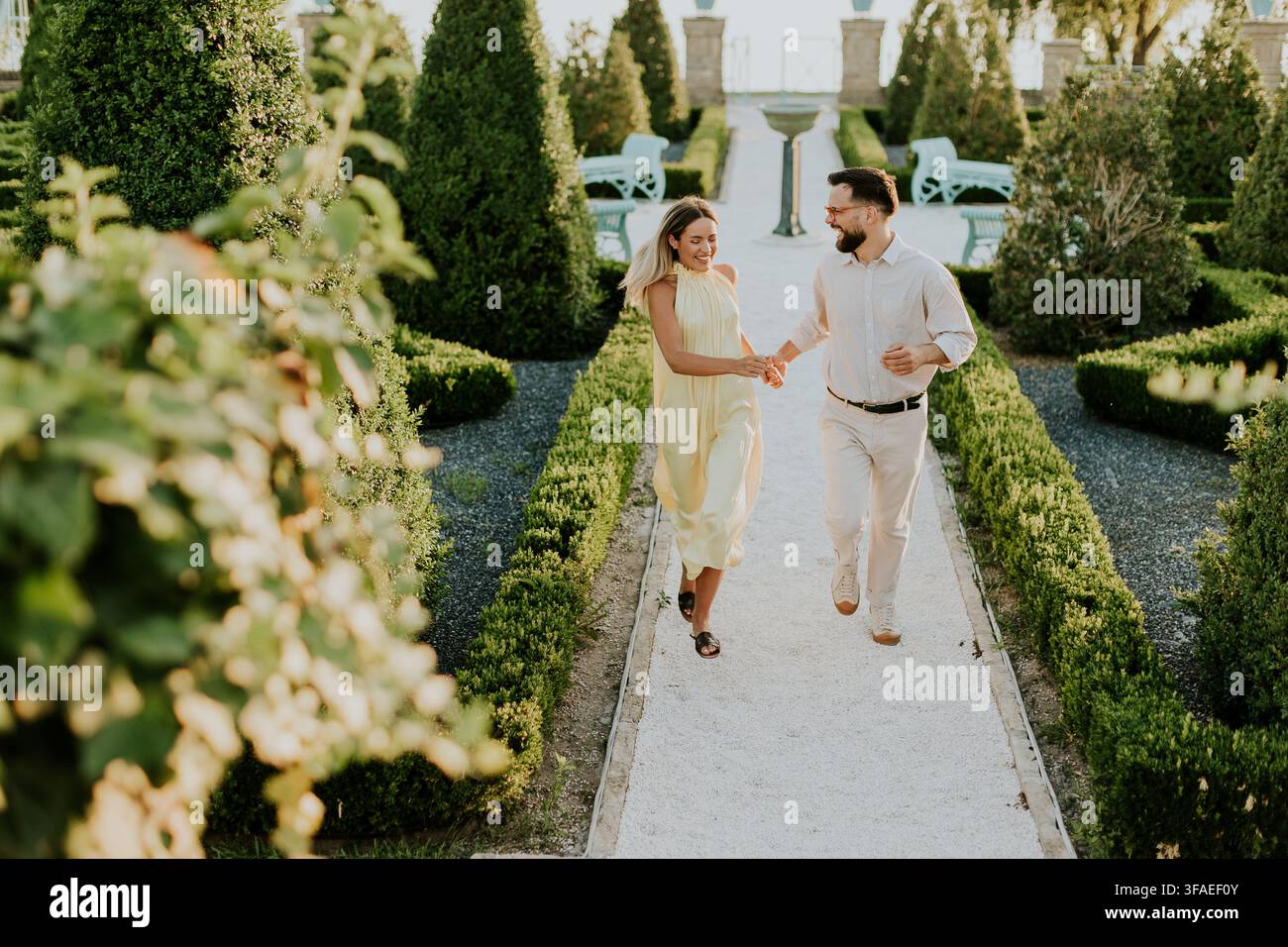 Ein Paar schlendert gemeinsam einen weißen Kiesweg in einem gepflegten Garten entlang. Sie halten sich an den Händen und lächeln, während sie den Moment unter der Sonne genießen. Stockfoto