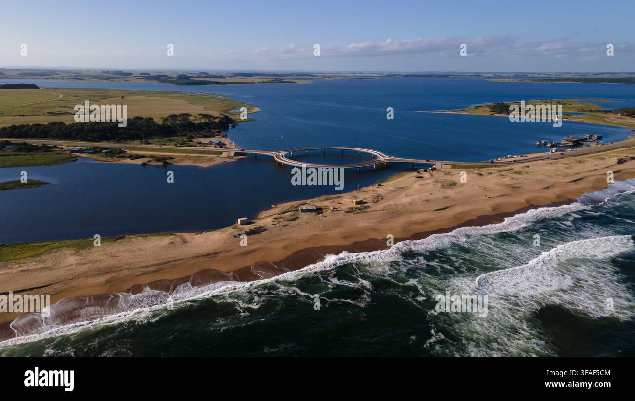 Luftaufnahme der ikonischen kreisförmigen Brücke über die Laguna Garzón, die Küstenbereiche in Maldonado, Uruguay, verbindet. Zeigt das lebendige blaue Wasser, sandigen B Stockfoto