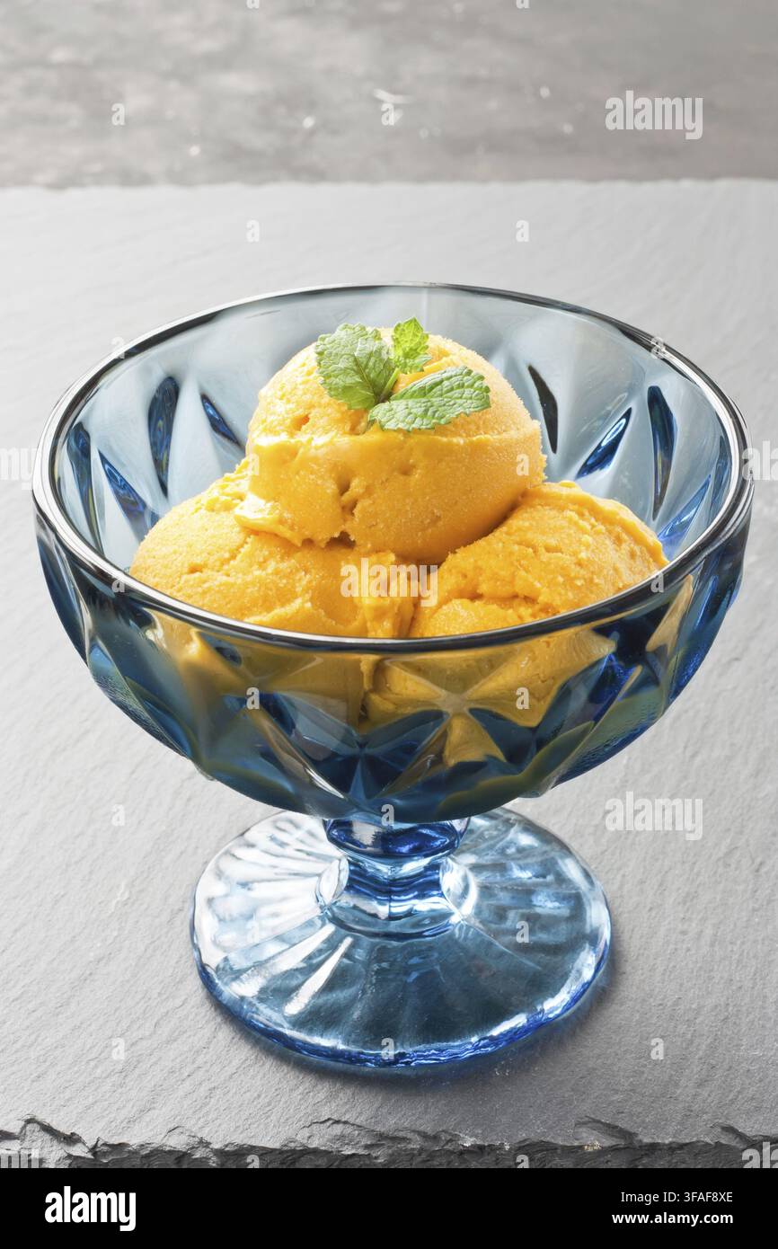 Mango-Eis mit Kokosmilch, verziert mit Minzblatt in blauer Glasschale auf dunkler Schieferplatte Stockfoto