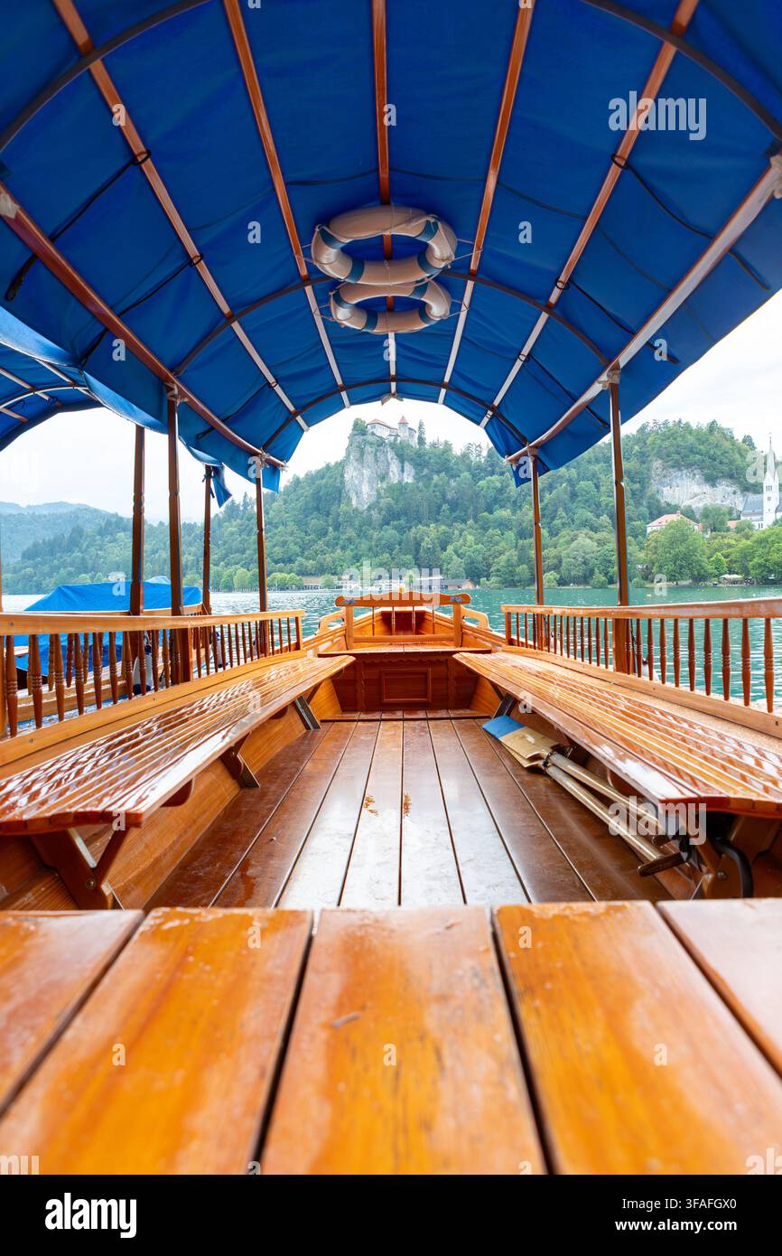In einem traditionellen hölzernen Pletna-Boot mit blauem Dach, nassen Bänken und Deck führt der Weg zum bewaldeten Ufer des Bled-Sees und zur Burg auf dem Felsen vor uns Stockfoto