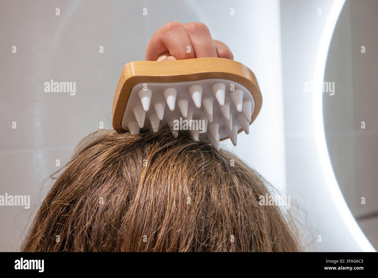 Ein Mädchen verwendet einen Kopfmassagegerät aus Holz und Silikon, um ihren Kopf in einem Badezimmer zu Hause zu massieren. Stockfoto