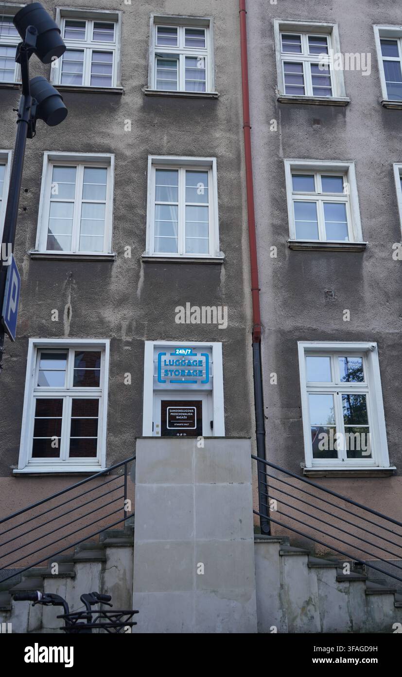 Danzig, Polen, 4. August 2026: Fassade eines alten Betonwohnhauses mit Außentreppen, die zu einem 24h/7-Gepäckaufbewahrungs-Eingang führen. Stockfoto
