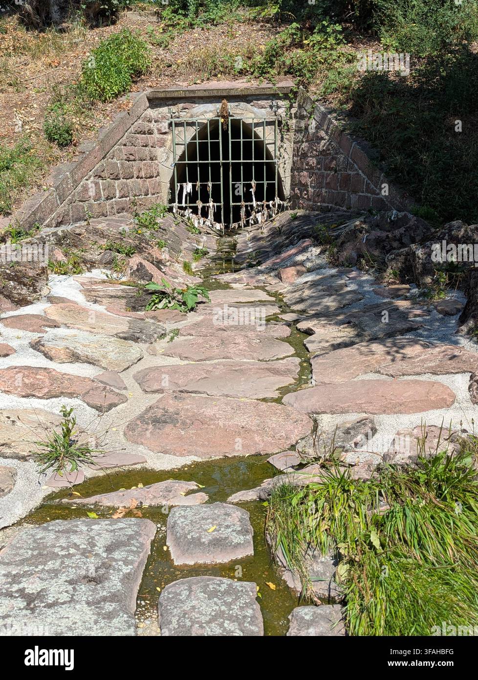Pforzheim Deutschland 2. August 2026 Ein ruhiger Steinbrunnen mit einem Metallgitter befindet sich in einer natürlichen Außenumgebung, umgeben von einem rustikalen Wasserlauf und r Stockfoto