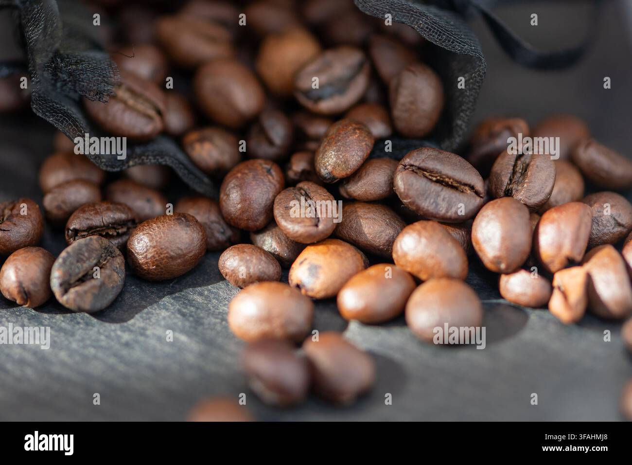 Geröstete Kaffeebohnen quellen aus einem schwarzen Stoffbeutel auf eine dunkle Oberfläche. Stockfoto