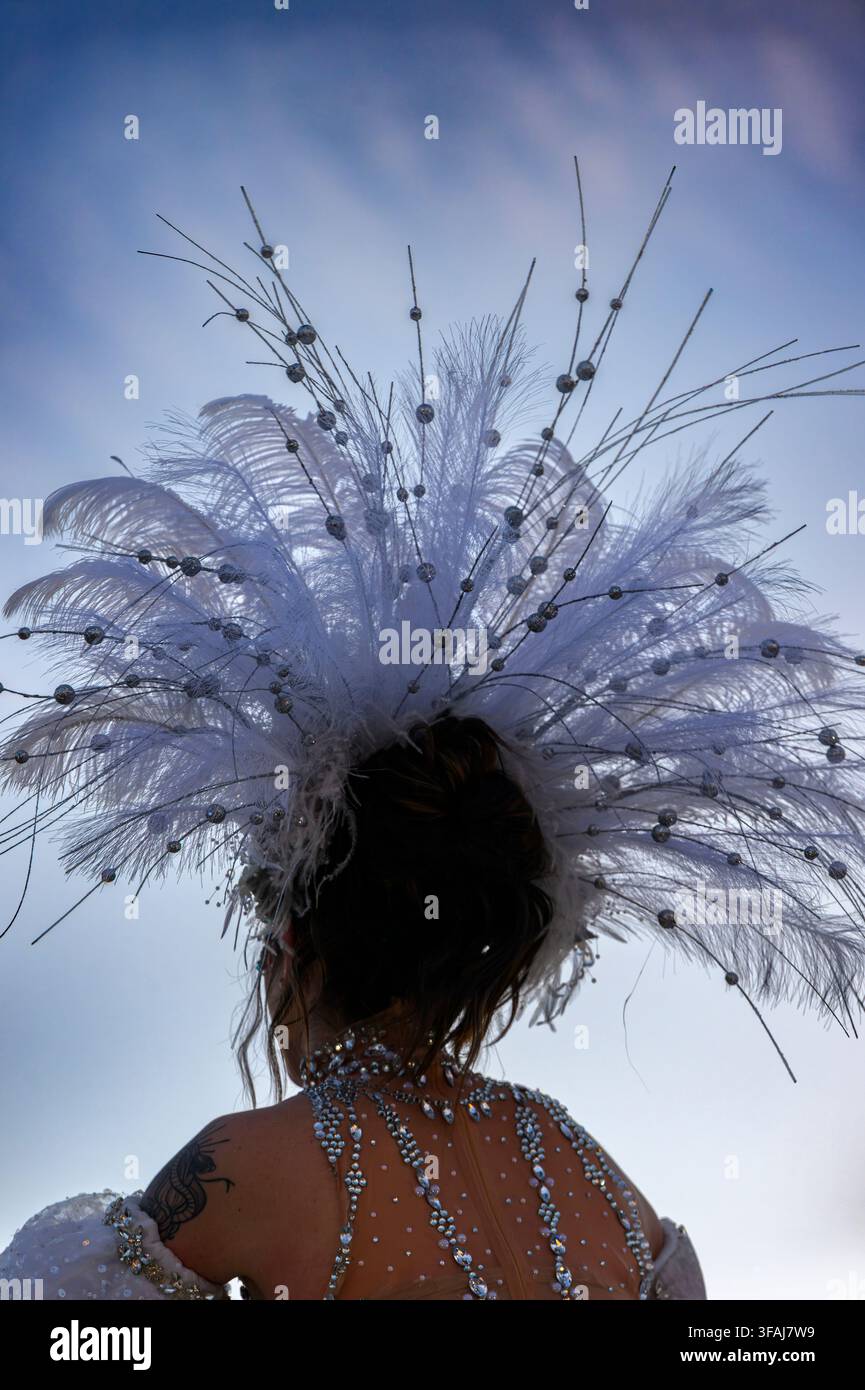 Frau trägt dramatische weiße Federhaube vor blauem Himmel. Federn sind weit gespreizt und fangen das Licht wunderschön ein. Rückansicht zeigt elegante Perlenstickerei auf s Stockfoto