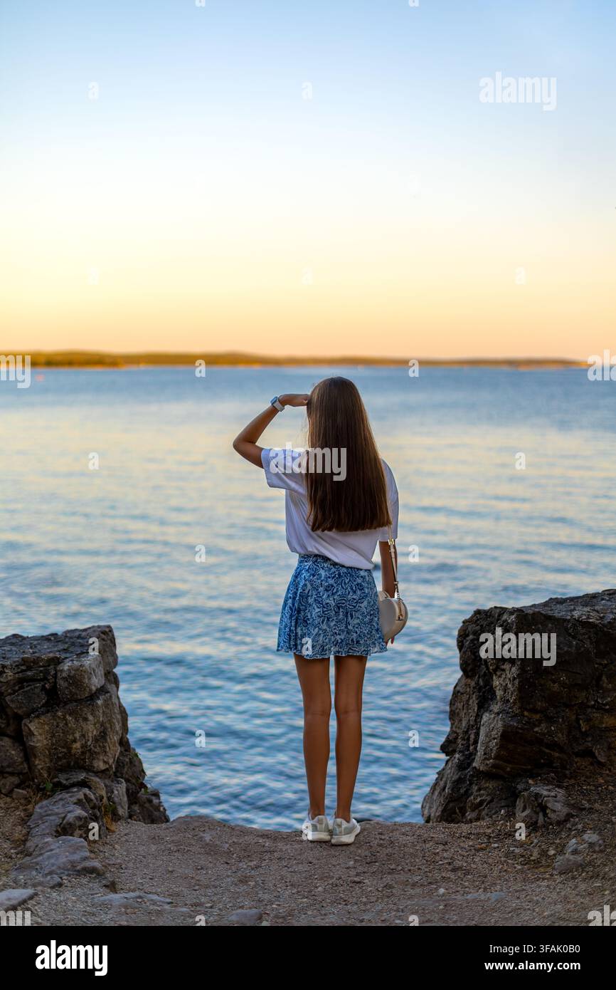 Frau blickt über ruhiges Wasser bei Sonnenuntergang. Frau steht an einem felsigen Ufer und blickt über ruhiges blaues Wasser in Richtung Horizont bei Sonnenuntergang. Stockfoto