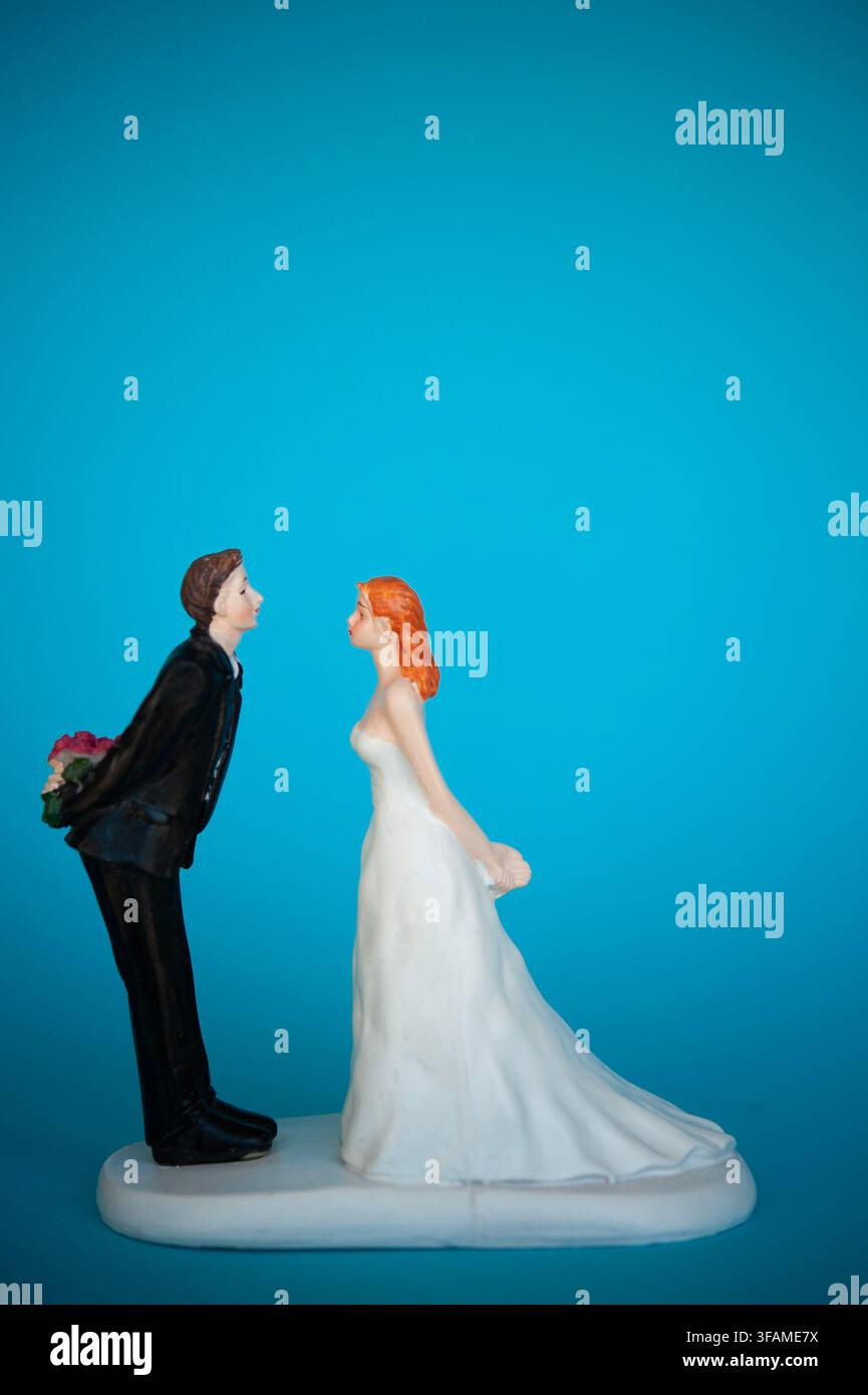 Hochzeitstorten-Topper einer Braut und eines Bräutigams, die sich gegenüberstehen, wobei der Bräutigam Blumen hinter seinem Rücken versteckt, auf einem hellblauen Hintergrund Stockfoto