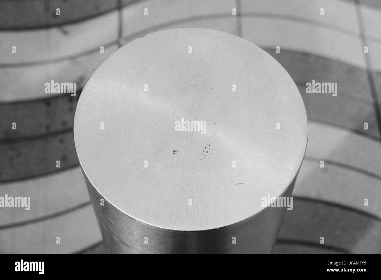 Oberansicht eines gebürsteten Metallzylinders vor gekrümmten Linien in einem gemusterten Pflaster. Stockfoto