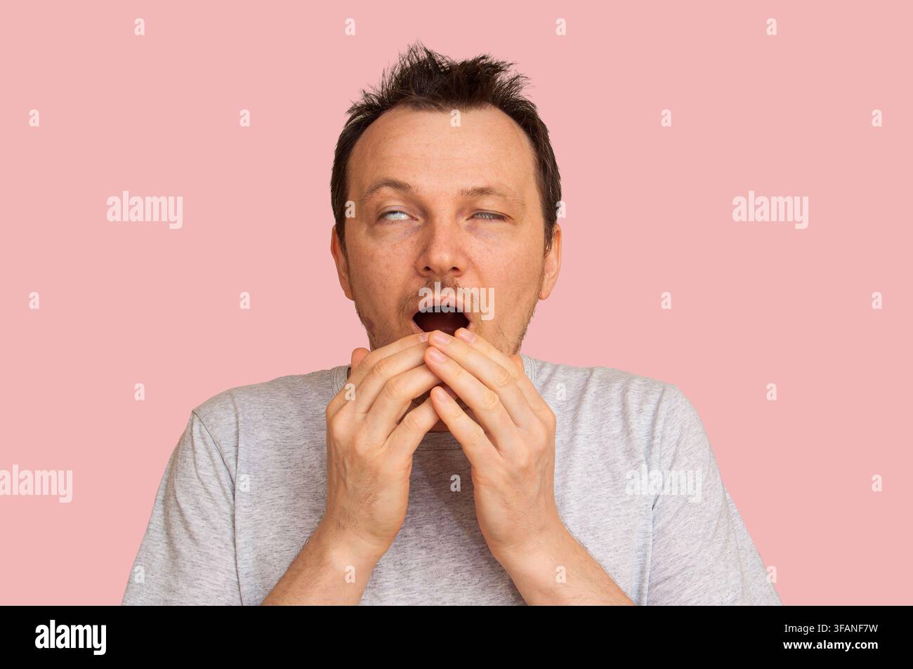 Ein müder Mann gähnt mit weit geöffnetem Mund und zurückgerollten Augen und bedeckt seinen Mund mit den Händen Stockfoto