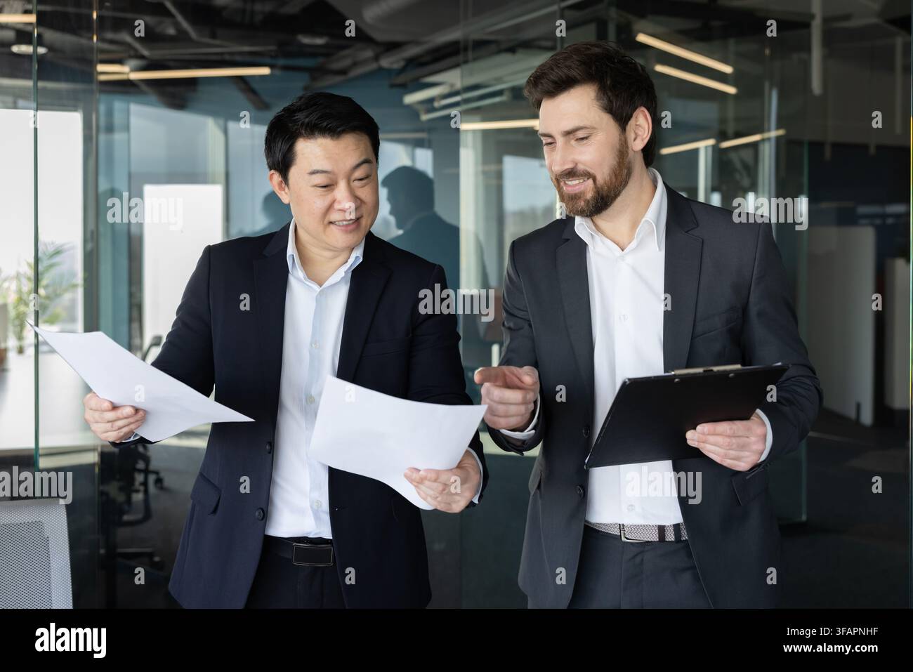 Zwei verschiedene Geschäftsleute in Anzügen, die in einem modernen Arbeitsumfeld während eines Unternehmertreffens an Dokumenten arbeiten, Finanzdaten überprüfen und wichtige Berichte besprechen Stockfoto