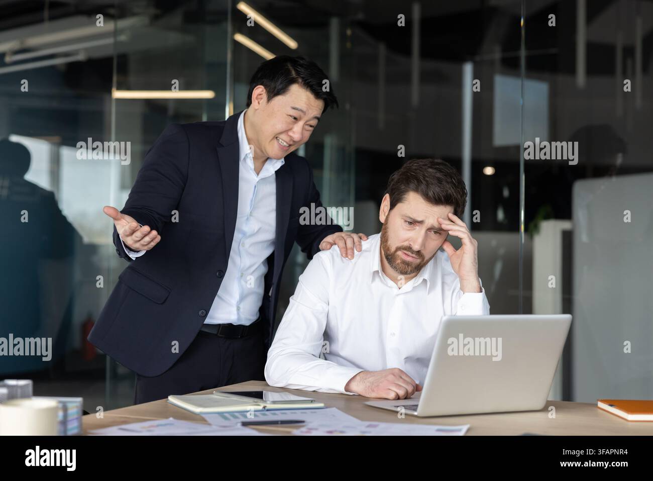 Asiatischer männlicher Chef, der über einem traurigen, gestressten kaukasischen männlichen Arbeiter an einem Laptop steht, zeigt Arbeitsplatzmobbing und Druck in einer modernen Büroumgebung Stockfoto