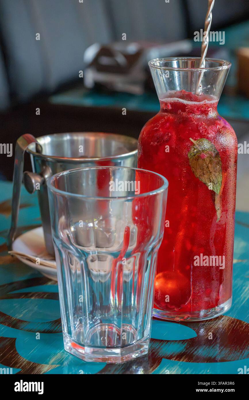 Ein erfrischendes Eistrink aus roten Beeren in einer Glaskaraffe wird neben einem leeren facettierten Glas und einem metallenen Eisbehälter auf einem blauen gemusterten Café-Tisch präsentiert Stockfoto