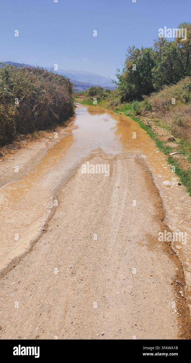 Überschwemmter unbefestigter Feldweg durch Wasserrohrbruch auf einer ländlichen Dorfstraße, umgeben von grünen Büschen, Stockfoto. Stockfoto