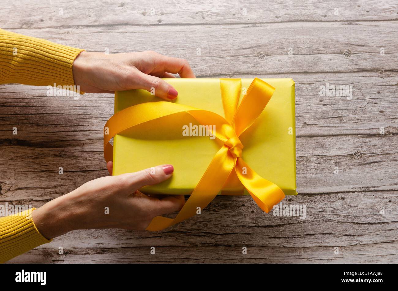 Frauenhände halten eine gelbe Geschenkschachtel mit gelber Schleife auf einem Holztisch. Stockfoto