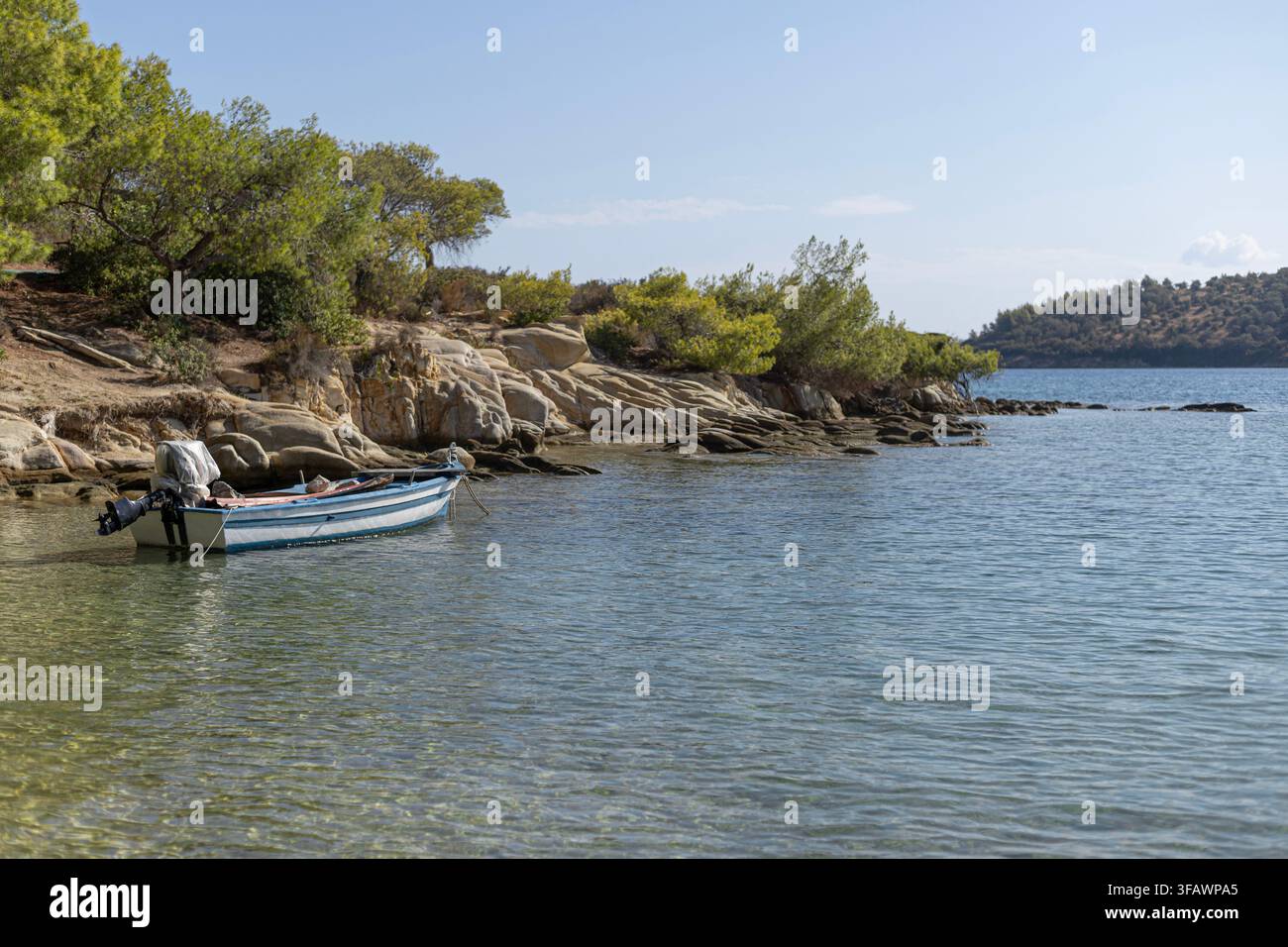 Ein kleines blau-weißes Motorboot liegt an einer felsigen Küste mit grünen Bäumen und Büschen unter einem klaren blauen Himmel. Scharfe Details des Wassers an Stockfoto