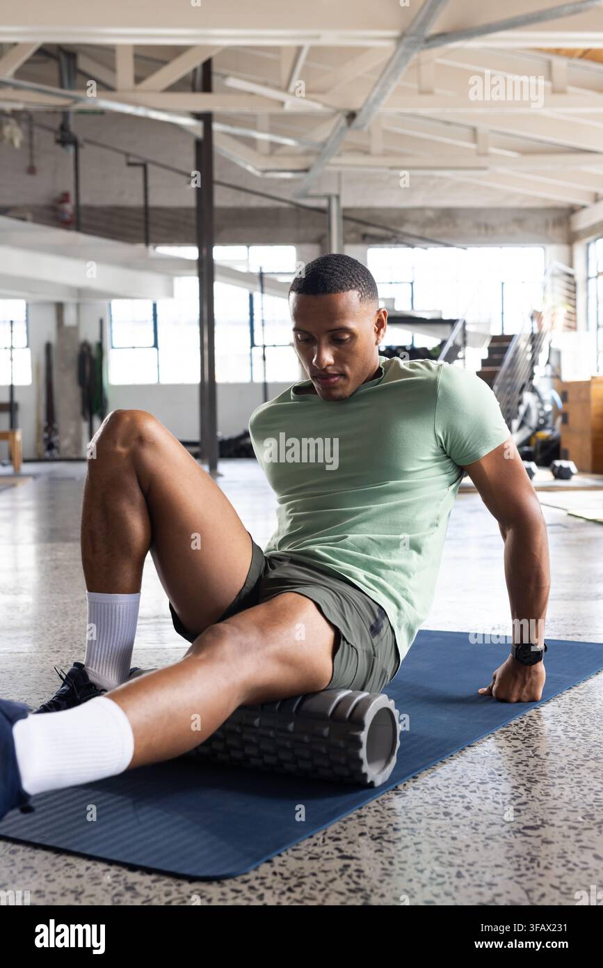 Erwachsener afroamerikanischer Mann sitzt auf einer Matte im Fitnessstudio und rollt auf einer Schaumstoffrolle, trägt ein grünes T-Shirt Stockfoto