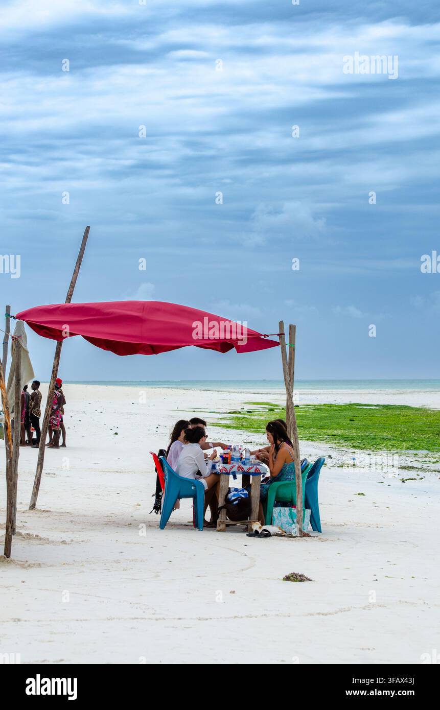 Menschen sitzen unter einem Strandschirm an einem weißen Sandstrand in Sansibar, Tansania Stockfoto