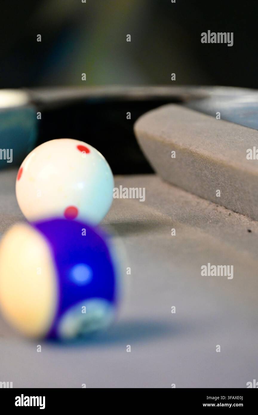 Eine vertikale Nahaufnahme mit scharfem Fokus auf dem weißen Spielball, der mit roten Punkten markiert ist, positioniert nahe dem Eckloch eines grauen Filz-Billardtisches, mit einem weich unscharfen lila gestreiften Ball im Vordergrund. Stockfoto