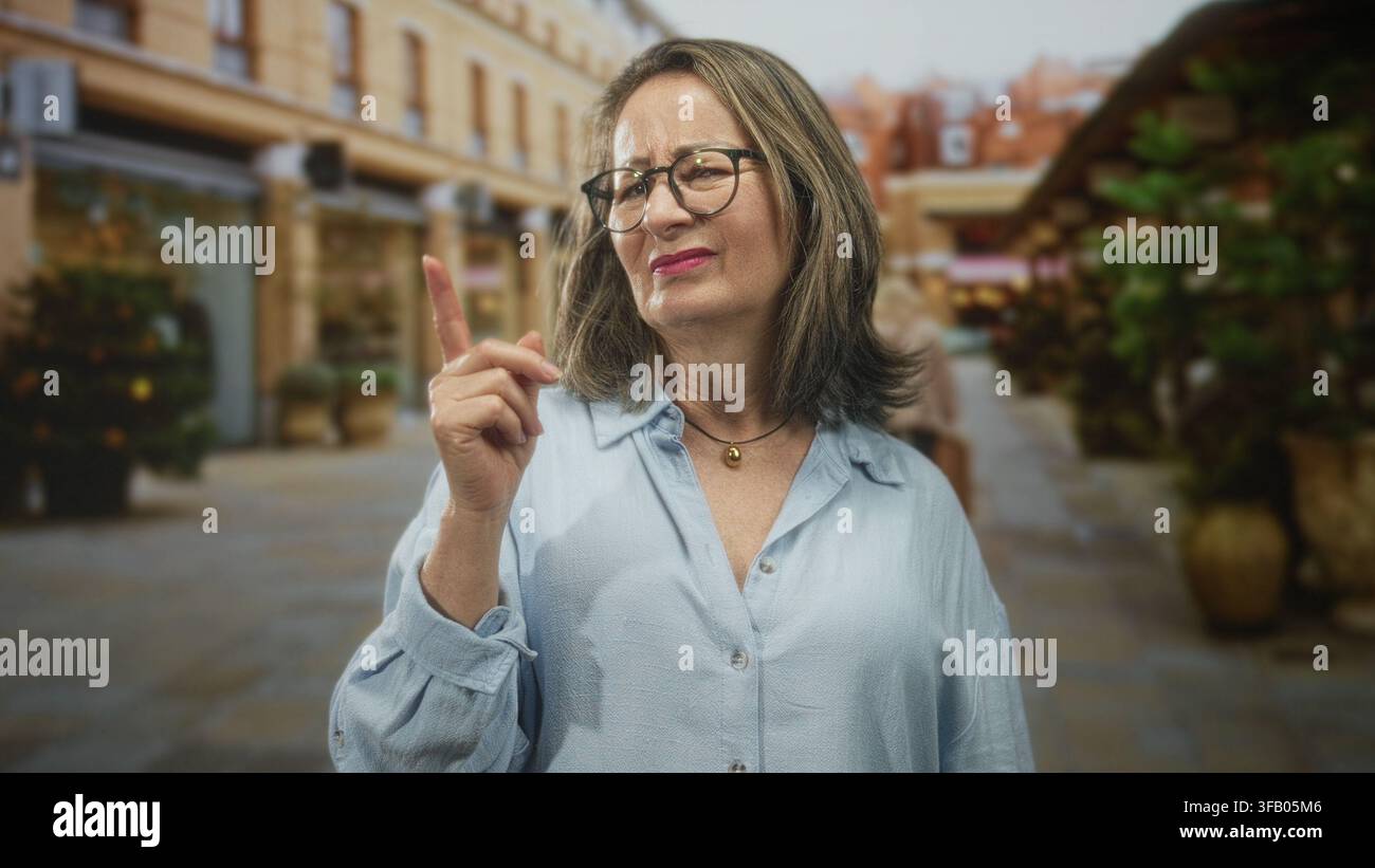 Hispanische Seniorin mit grauem Haar und Brille zeigt mit dem Zeigefinger und blinzelt, während sie in einer Straße steht, die von Geschäften und Pflanzern gesäumt ist; Missbilligung Stockfoto