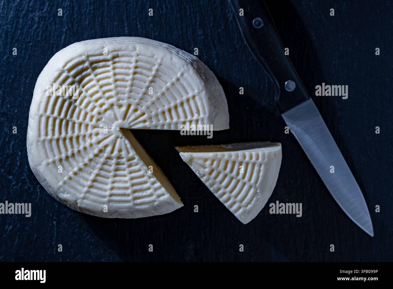 Frischer traditioneller hausgemachter Käse mit Messer auf dunklem Hintergrund, Nahaufnahme. Oberansicht einer runden Käsekugel mit abgeschnittenem Stück und Messer auf Schwarz Stockfoto