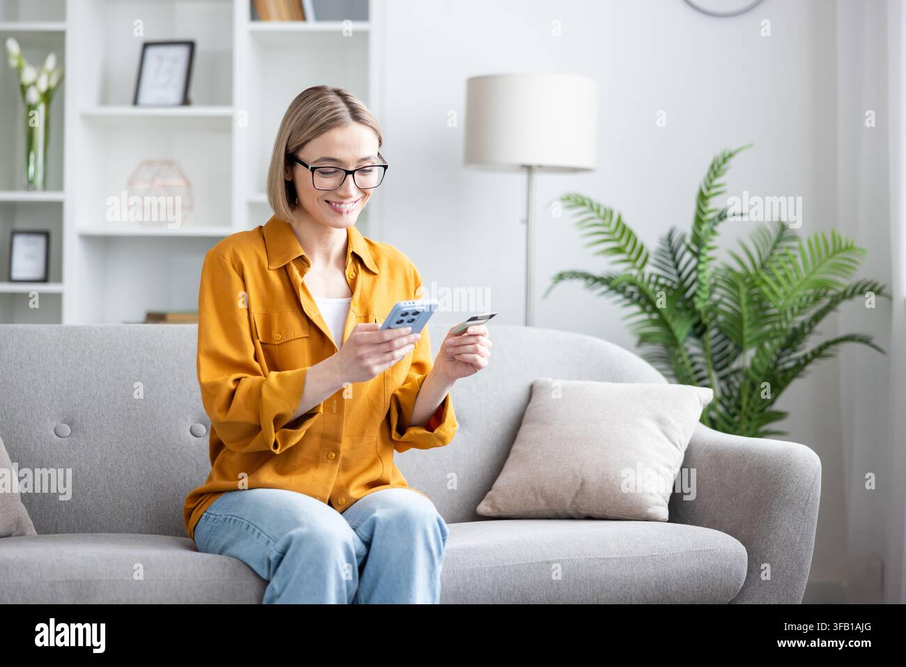 Junge Frau auf einem Sofa zu Hause, lächelt, während sie ihr Smartphone benutzt, um Kreditkartendaten für eine sichere Online-Zahlung einzugeben, während sie in legerer Kleidung entspannt Stockfoto