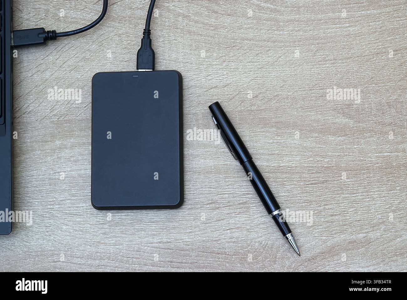 Flachlayout-Perspektive einer schwarzen tragbaren externen Festplatte, die über ein USB-Kabel an einen Laptop angeschlossen ist, gepaart mit einem raffinierten schwarz-silbernen Stift Stockfoto