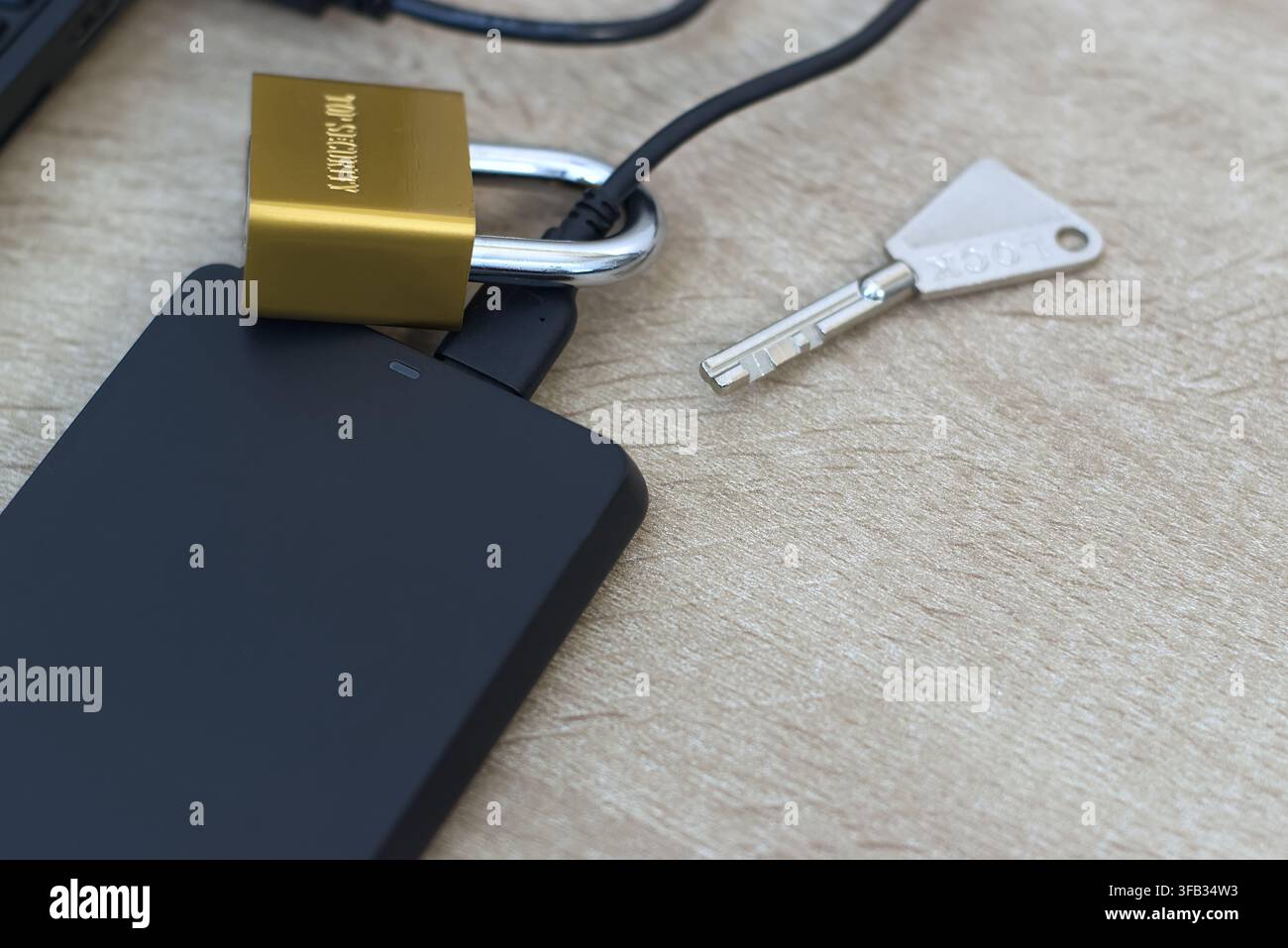 Nahaufnahme eines kleinen Messingschlosses, das physisch um das USB-Kabel einer schwarzen externen Festplatte gesichert ist. Laptop-Tastatur seitlich sichtbar. Co Stockfoto
