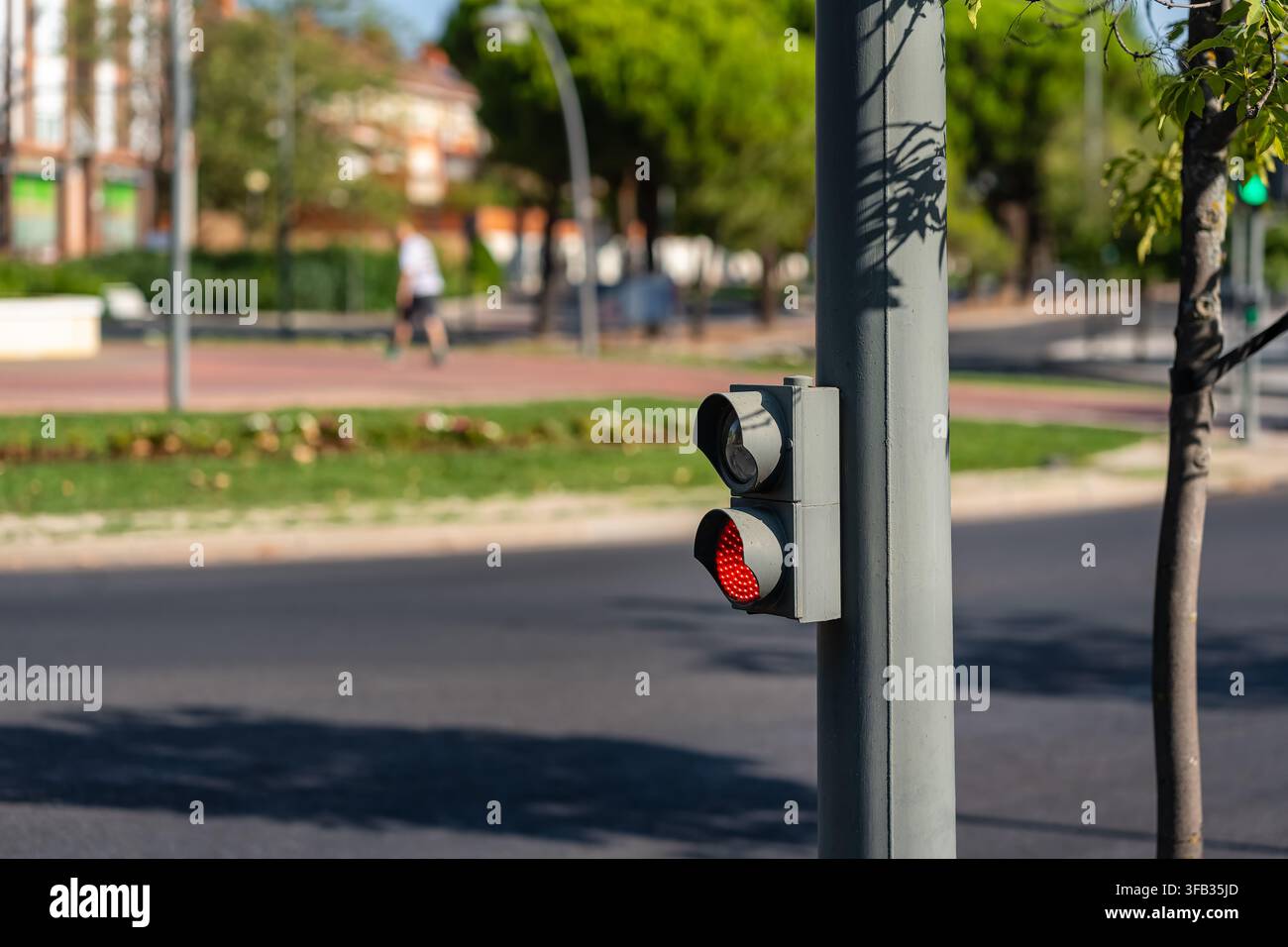 Kleine rote Ampel neben einer städtischen Straße mit Bäumen und einem verschwommenen Läufer im Hintergrund Stockfoto