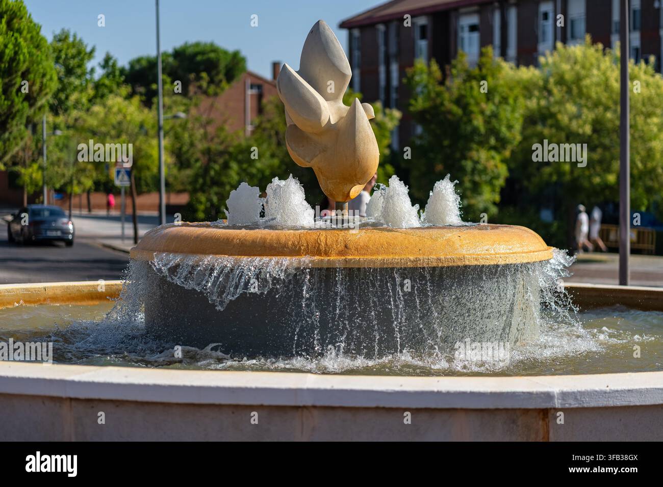 Abstrakte Steinskulptur über einem fließenden städtischen Brunnen an einer sonnigen, von Bäumen gesäumten Stadtstraße Stockfoto