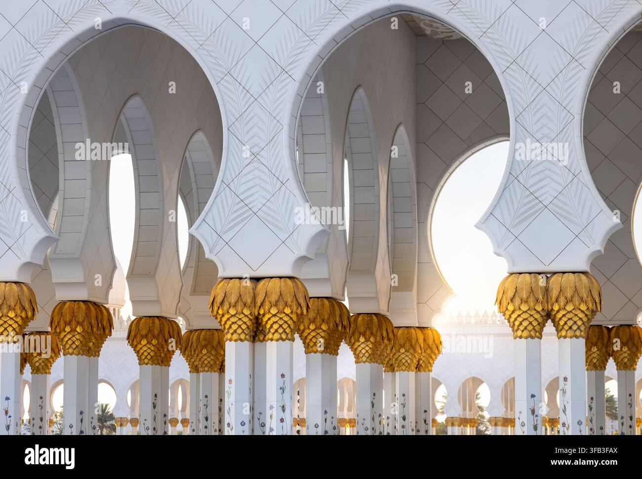Elegante weiße Bögen und goldene Palmkapitelle umrahmen den sonnenbeschienenen Innenhof der Scheich-Zayed-Großmoschee in Abu Dhabi und präsentieren aufwendige islamische Architektur Stockfoto