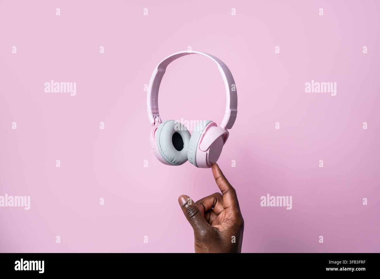 Nahaufnahme einer schwarzen Hand, die rosa Over-Ear-Kopfhörer auf einem Finger balanciert, vor einem weichen pastellrosa Hintergrund, wodurch ein verspieltes minimales Audiovisuales entsteht Stockfoto