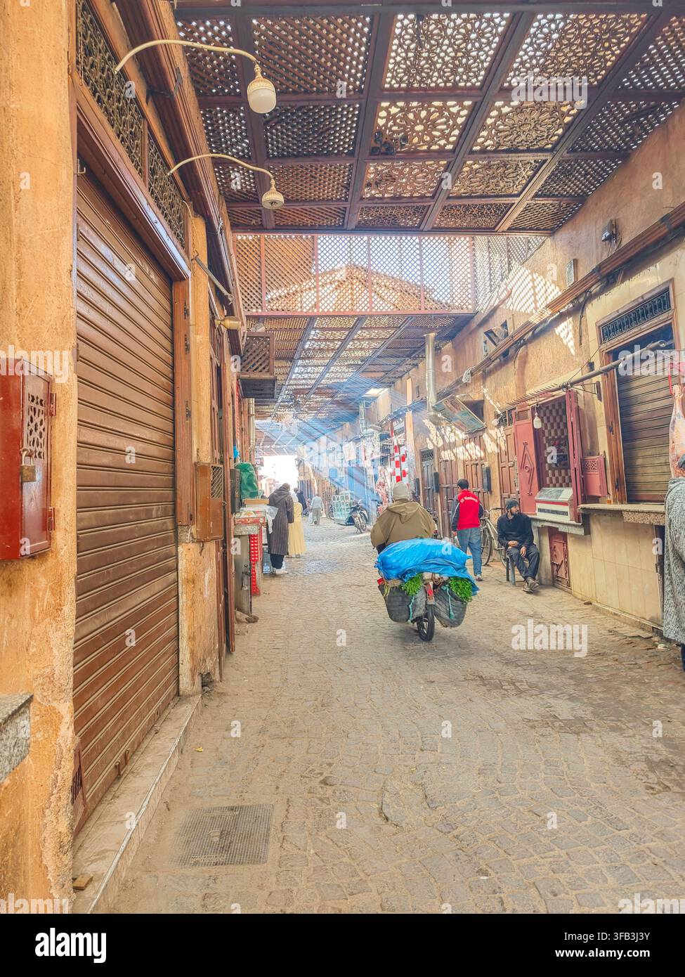 Traditionelle Marktgasse in der Medina von Marrakesch mit einem überdachten Holzlattichdach, lokalen Geschäften und einem Händler, der Waren mit dem Fahrrad transportiert. Stockfoto