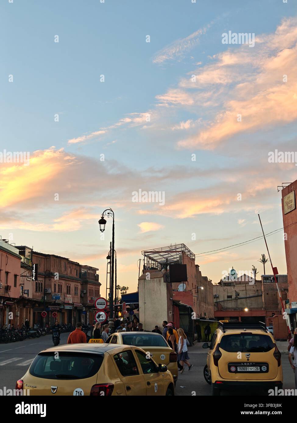 Straßenszene in Marrakech bei Sonnenuntergang mit gelben Taxis, Fußgängern, Motorrädern und traditionellen marokkanischen Gebäuden, Marokko. Stockfoto