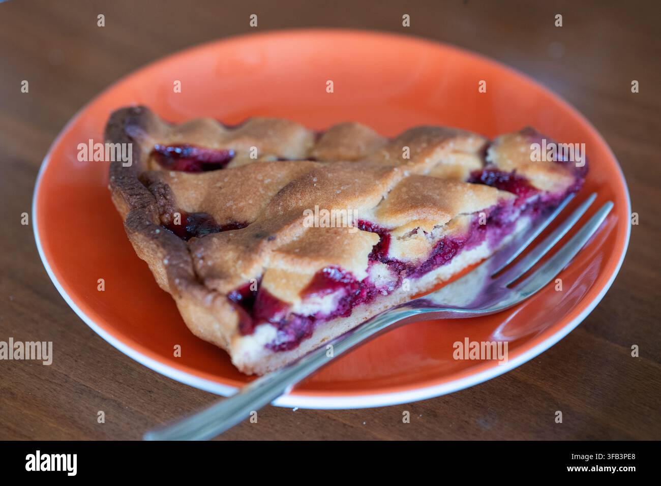 Eine Nahaufnahme eines frisch gebackenen Stücks hausgemachter Kirsch-Kästchenkuchen, serviert auf einem orangefarbenen Teller mit einer silbernen Gabel, vor einem sanft verschwommenen Woo Stockfoto
