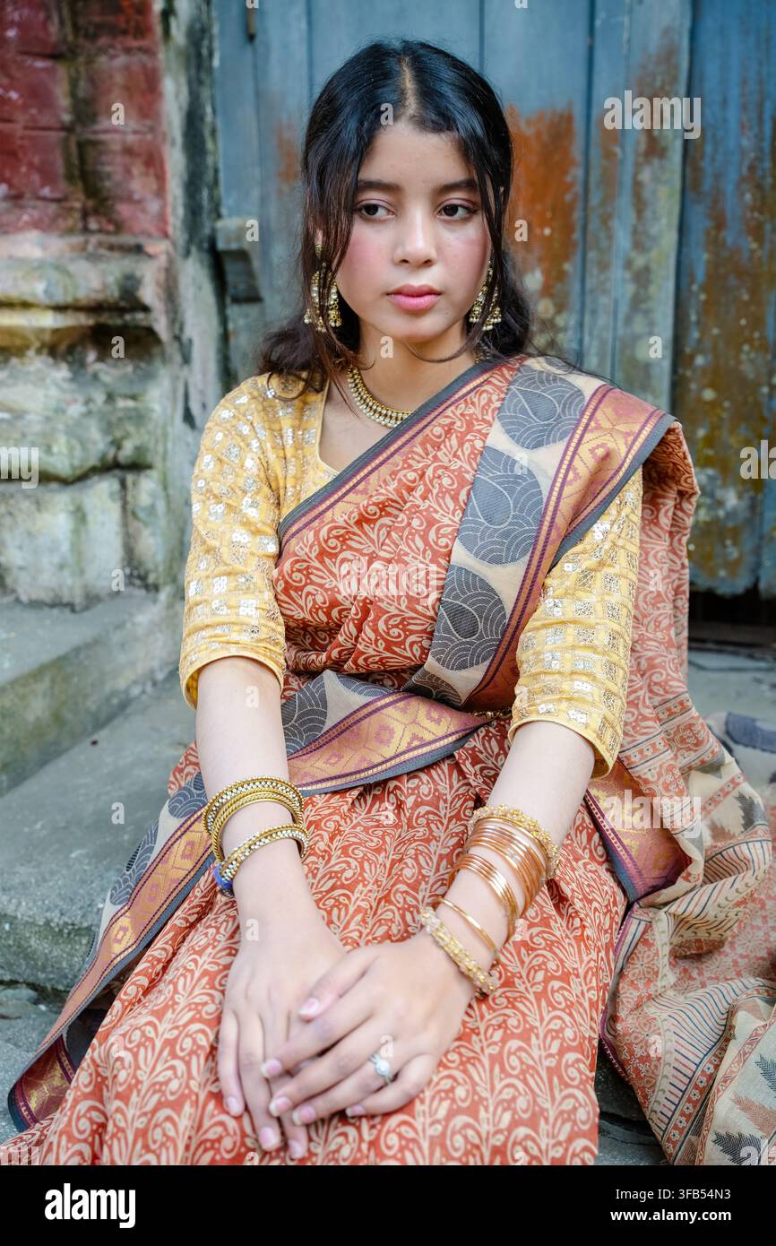 Eine Frau sitzt im Freien und trägt einen traditionellen Saree mit aufwendigen Mustern und Goldschmuck und posiert vor einem verwitterten Ziegelwandhintergrund. Stockfoto