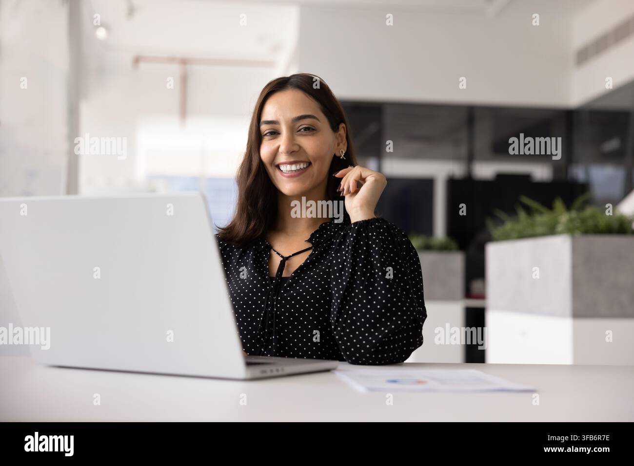 Selbstbewusste Geschäftsfrau lächelt in die Kamera, während sie an einem Schreibtisch mit Laptop sitzt Stockfoto
