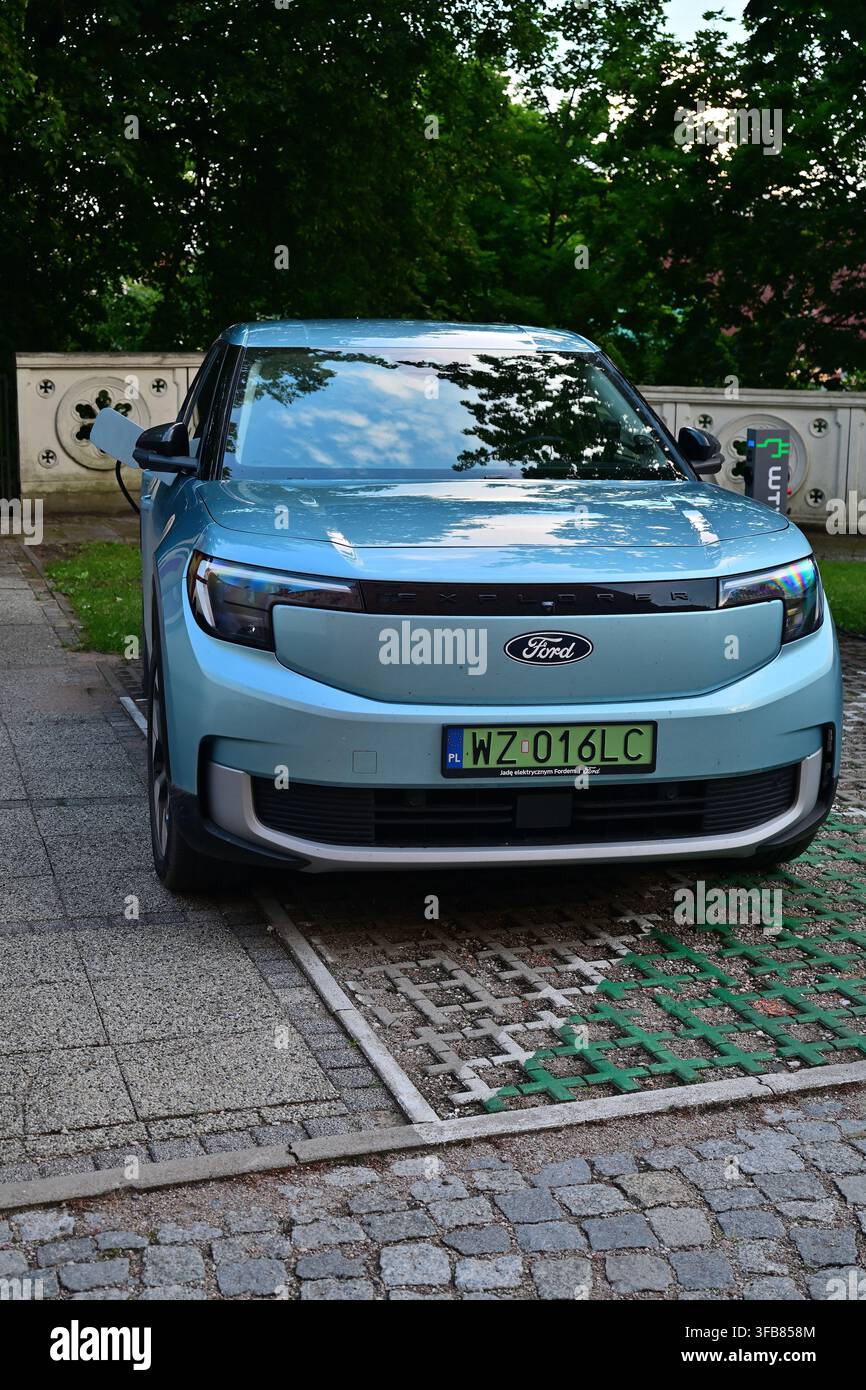 Blauer Ford Explorer Elektroauto lädt an einem städtischen Parkplatz auf, umgeben von Bäumen und Spiegelung. Stockfoto