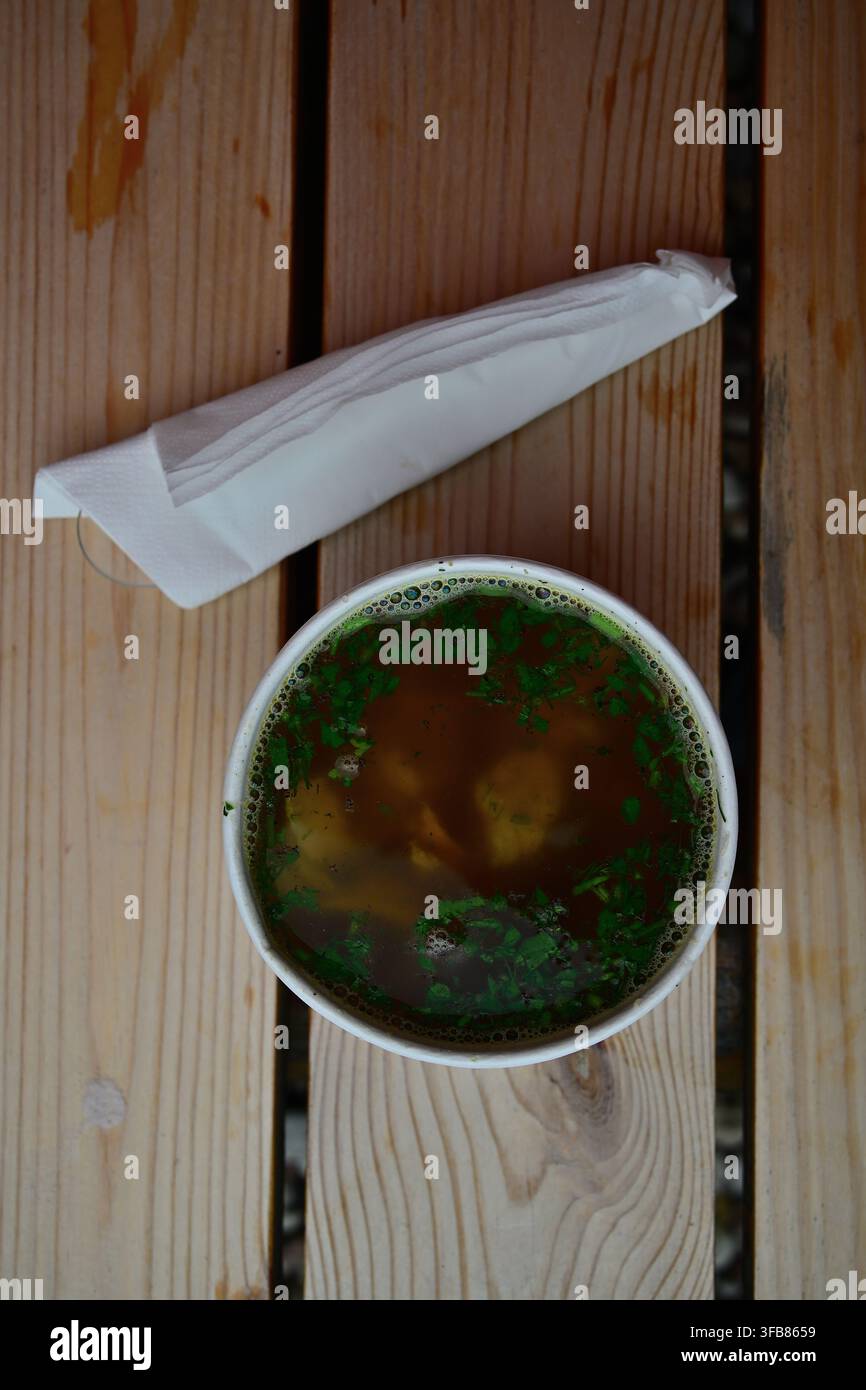 Klare Brühesuppe mit Fischstücken in einer Papierschale mit Kräutern auf einem Holztisch. Stockfoto