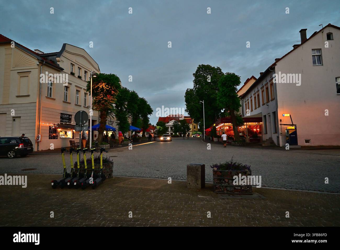 Abendlicher Stadtplatz mit Cafés, Straßenlaternen und geparkten Elektro-Scootern in der historischen polnischen Stadt – der masurischen Stadt Mikołajki. Stockfoto