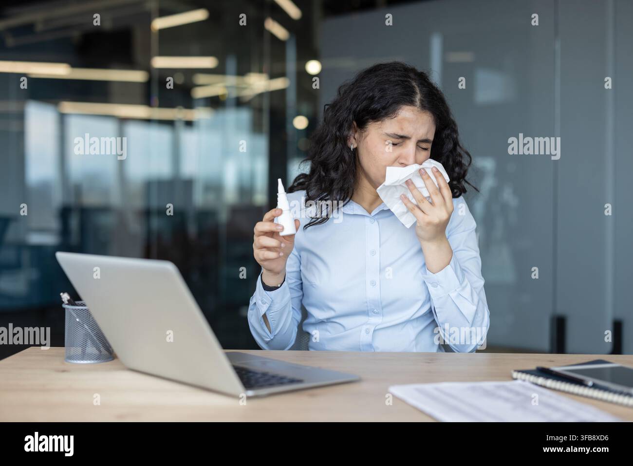 Junge Geschäftsfrau, die unter Erkältungs- oder Allergiesymptomen leidet, schnäuzt sich mit einem Taschentuch die Nase und hält ein Nasenspray, während sie an ihrem Schreibtisch im Büro arbeitet Stockfoto