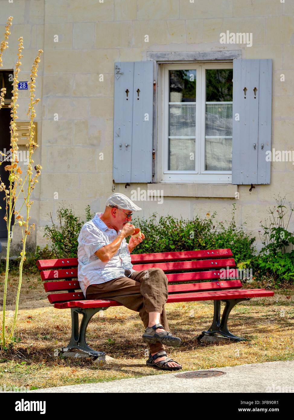 Älterer Mann isst ein Eis und sitzt auf einer langen Bank auf dem Platz in der Stadt - Beaulieu-lès-Loches, Indre-et-Loire (37), Frankreich. Stockfoto