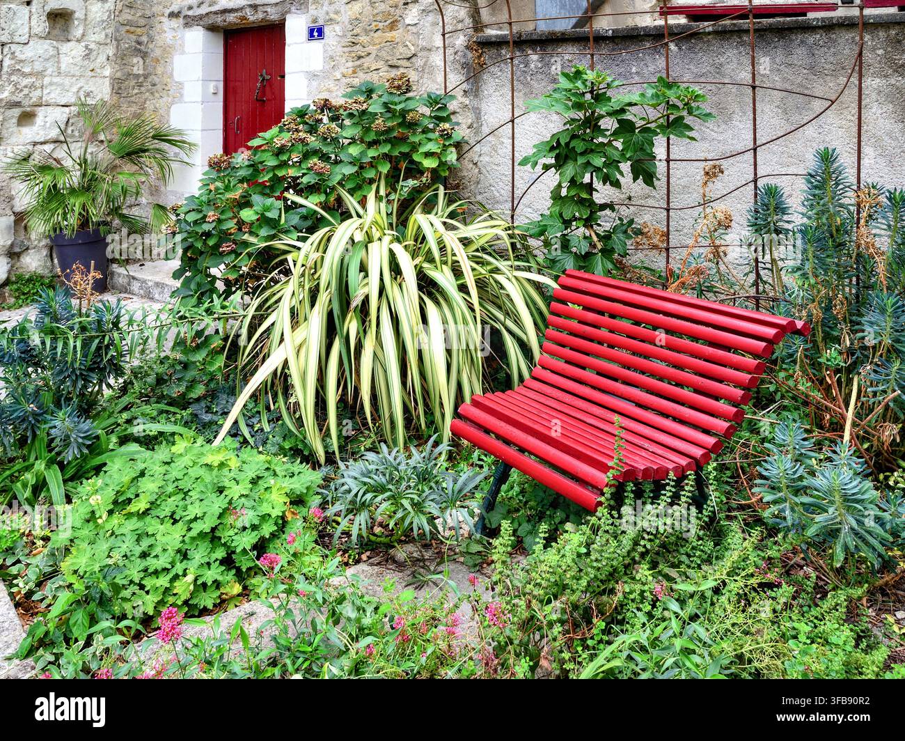 Rot gestuhltes kleines Gartensitzgelegenheit inmitten eines dekorativen Blumengartens - Beaulieu-lès-Loches, Indre-et-Loire (37), Frankreich. Stockfoto