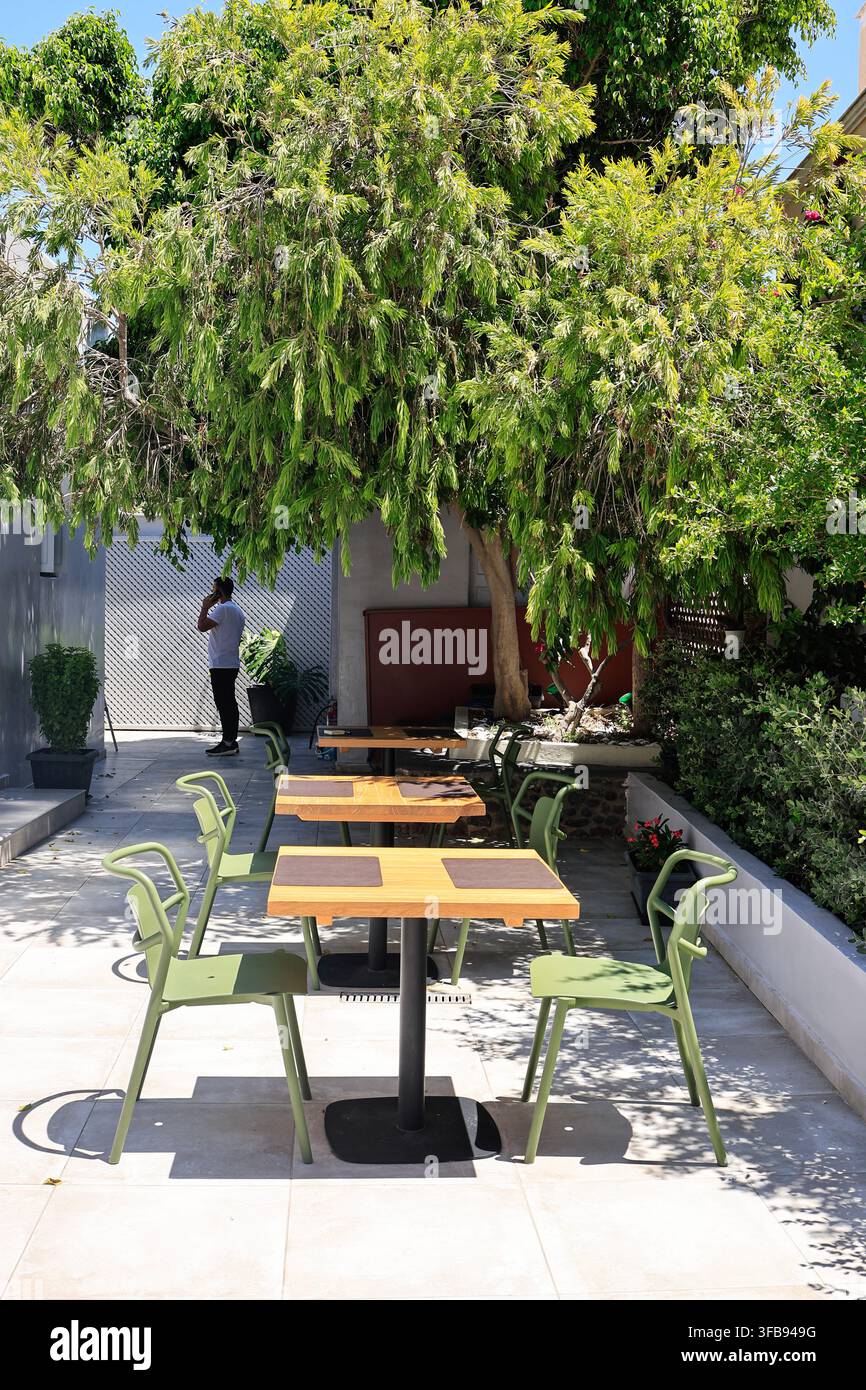 Oia, Santorin, Griechenland - 4. August 2026: Leere Café-Tische und grüne Stühle unter einem schattigen Baum Stockfoto