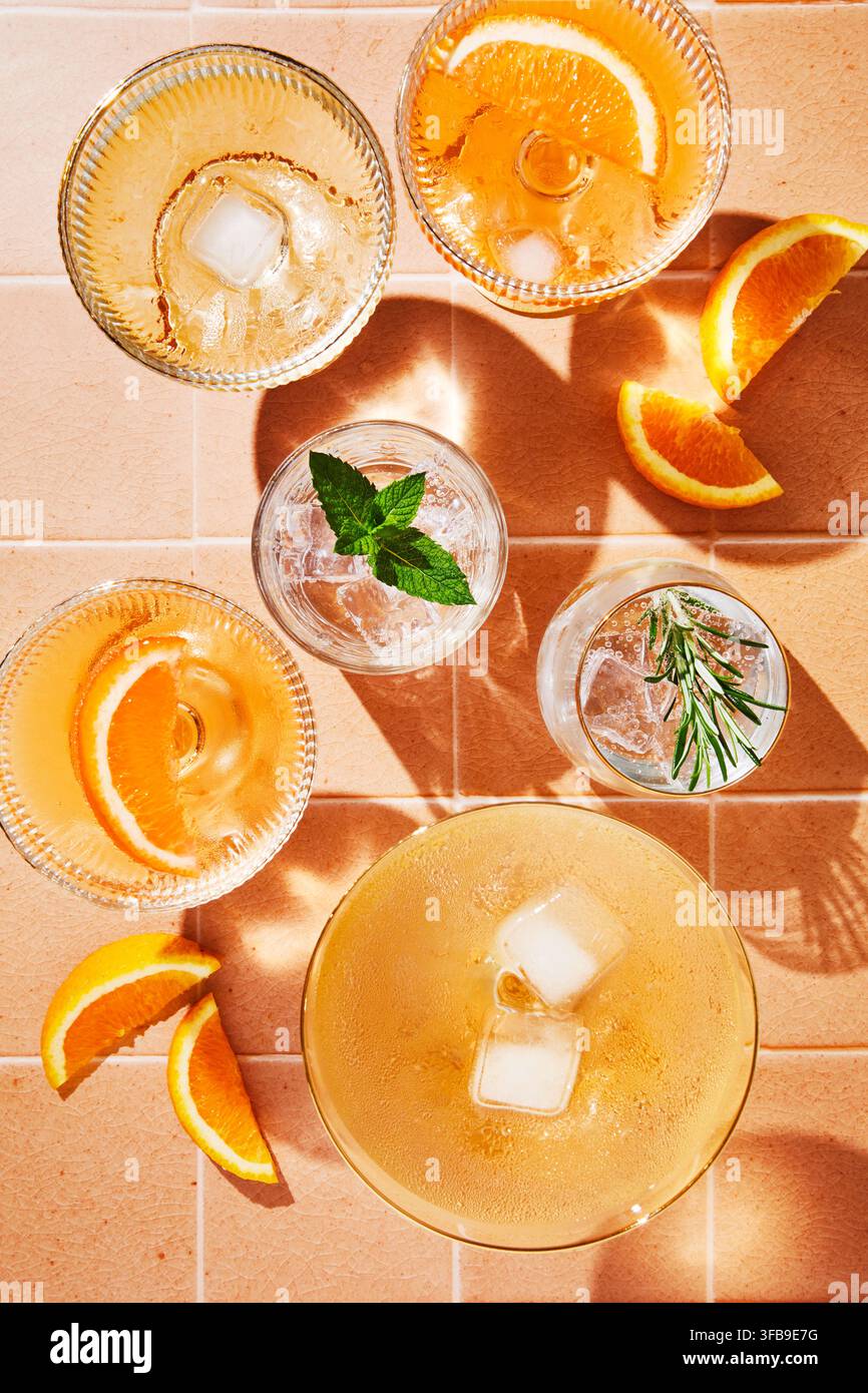 Verschiedene Gläser gefüllt mit erfrischenden Sommergetränken auf gelben Fliesen. Orangenscheiben, Minzblätter, Rosmarin und Eiswürfel. Sonnenlicht. Stockfoto