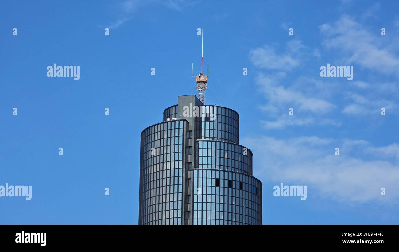 Der obere Teil eines markanten, gestuften Hochhauses aus dunklem Glas mit einem hohen orange-weißen Funkkommunikationsmast vor einem blauen Himmel Stockfoto