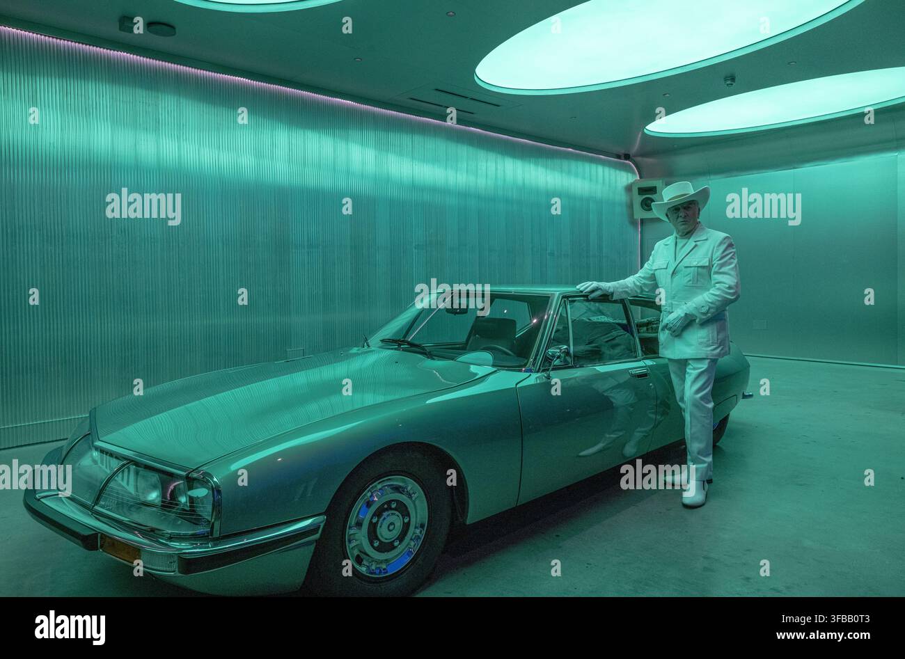 posiert in einem komplett weißen Westernanzug neben seinem klassischen Citroën SM Grand Tourer Coupé von 1971 in einem futuristischen Raum unter grünem Neonlicht. Stockfoto