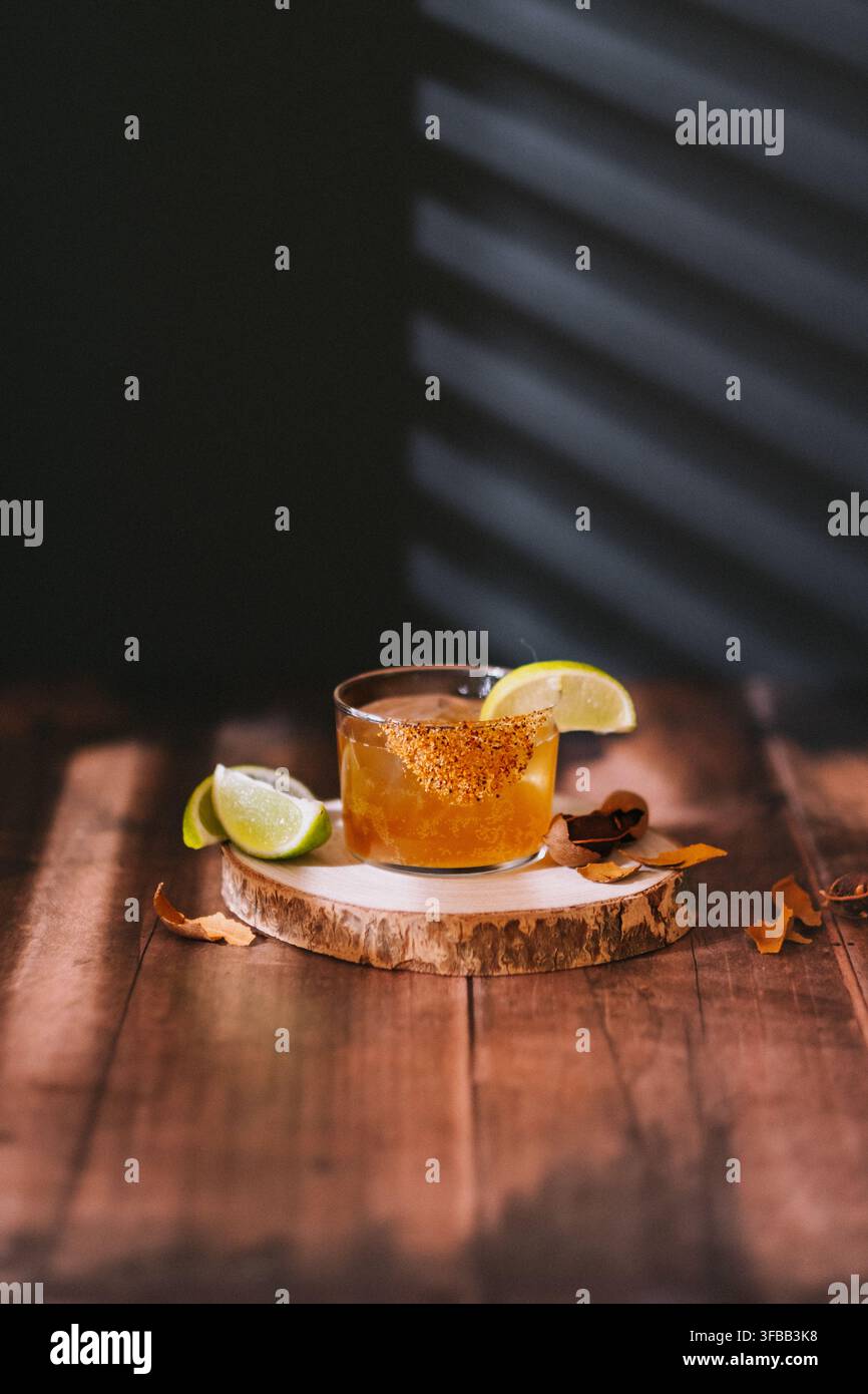 Mezcal-Cocktail mit einem Chilischalenrand und Limettenspaltern auf einem Holzbrett auf einem rustikalen Tisch mit Fensterschattierungen Stockfoto