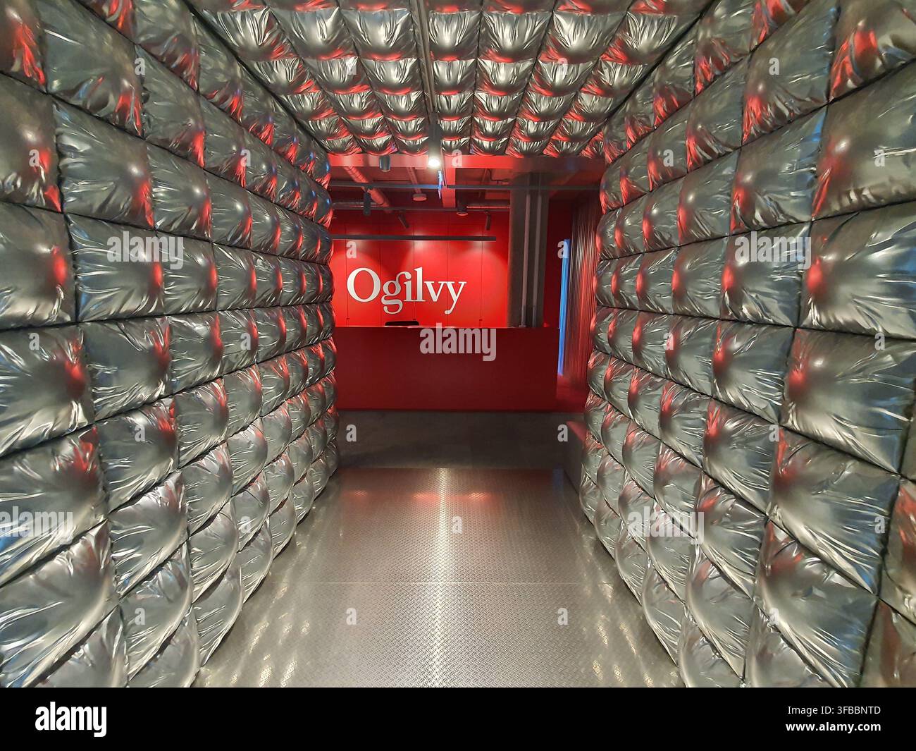 Ein futuristischer Korridor führt zu einer hellroten Wand mit dem weißen „Ogilvy“-Logo, das den Raum als Eigentum der Werbeagentur kennzeichnet. Stockfoto