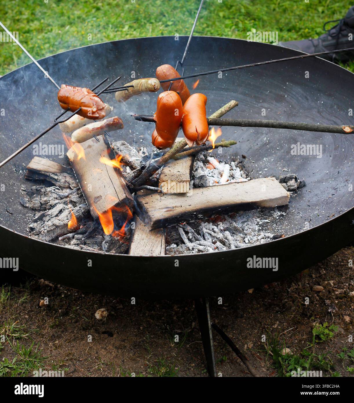 Tragbarer Feuerkorb mit bratenden Würsten über offenem Feuer während eines Outdoor-Grillfestes und Campingtreffens Stockfoto